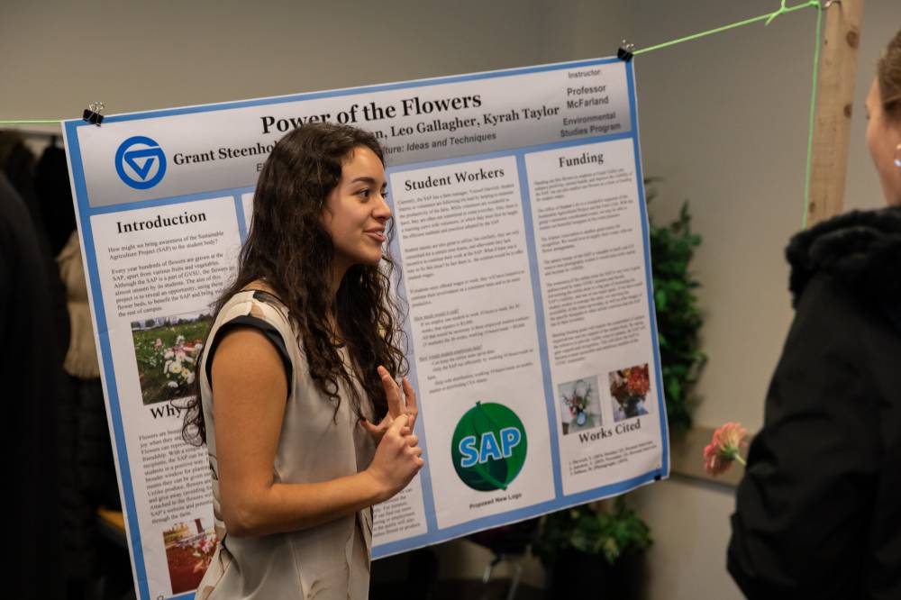 A student smiles while speaking to guests about her poster regarding flowers
